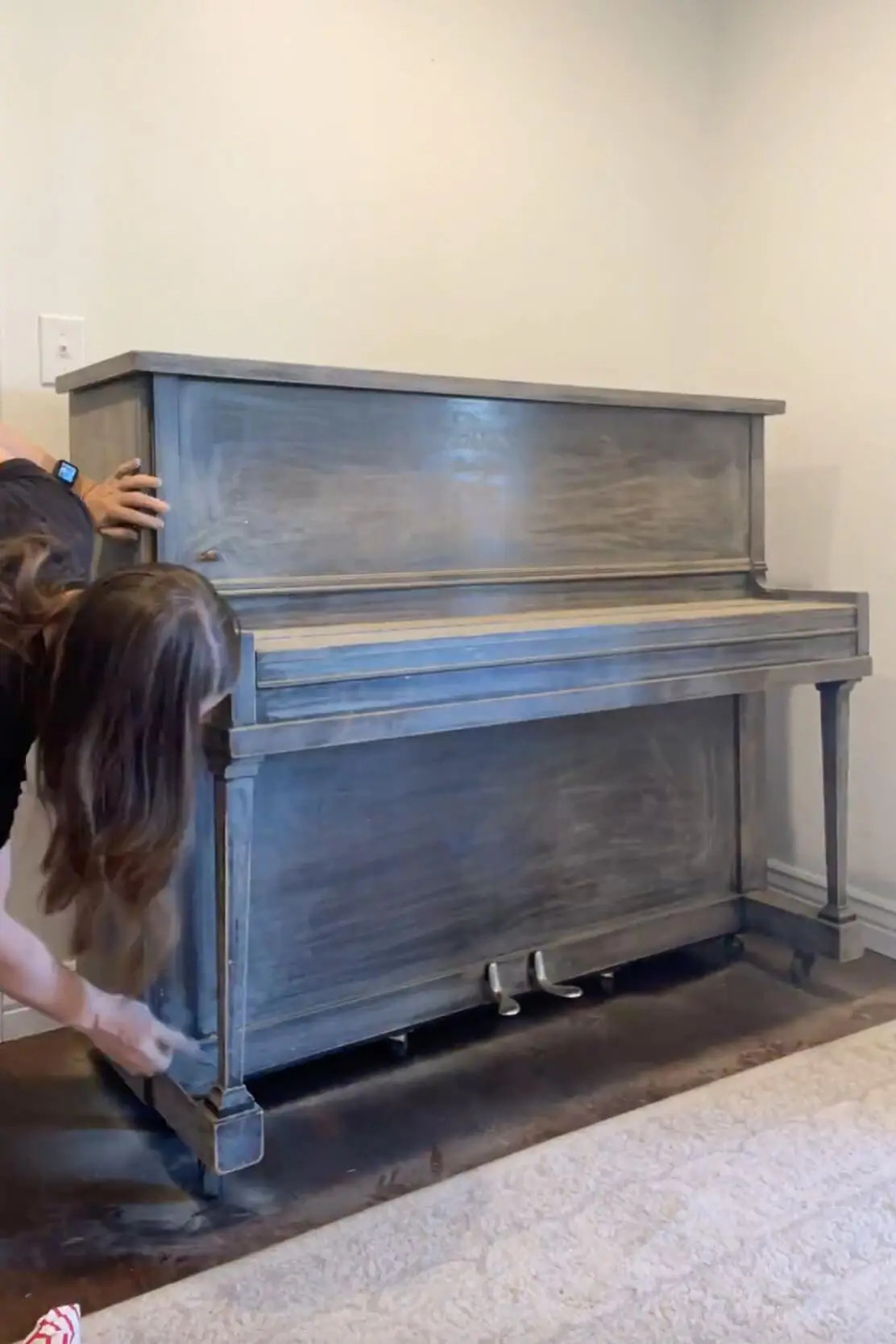 Woman and child sanding down a dark brown upright piano, removing the old finish and preparing the surface for a fresh coat of paint.